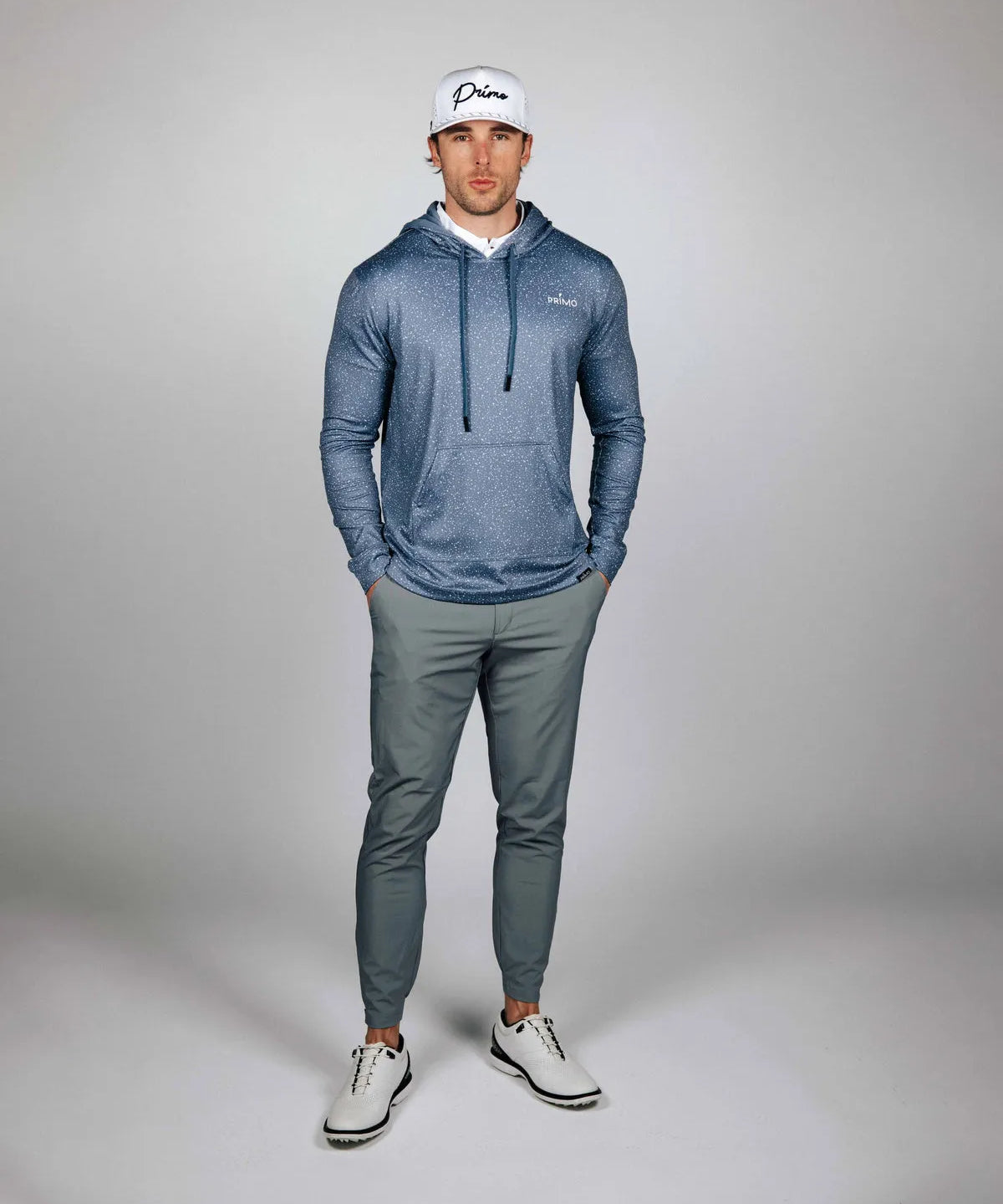 Man wearing a blue hoodie and gray pants with a white cap on a gray background