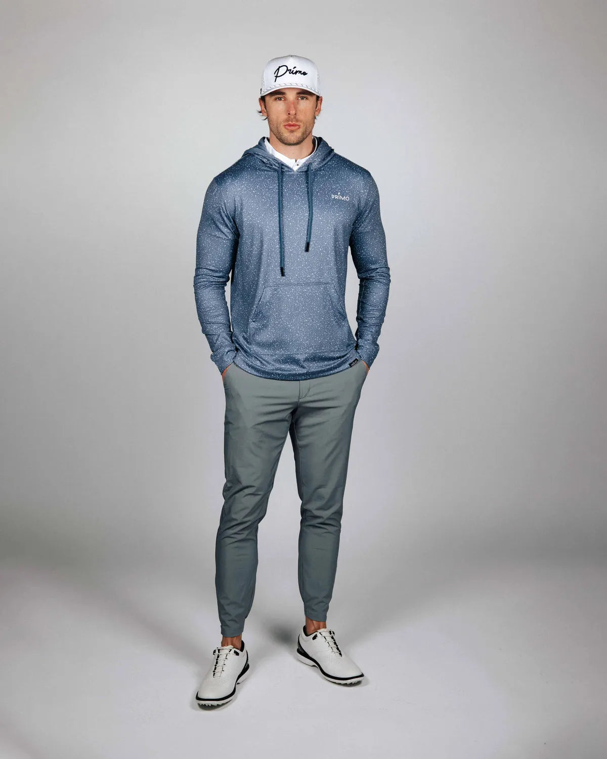 Man wearing a blue hoodie and gray pants with a white cap on a gray background