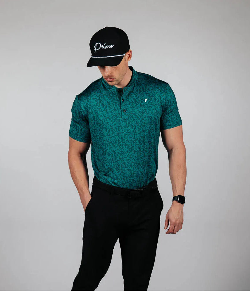 Man wearing a green patterned shirt, black pants, and a black cap on a gray background