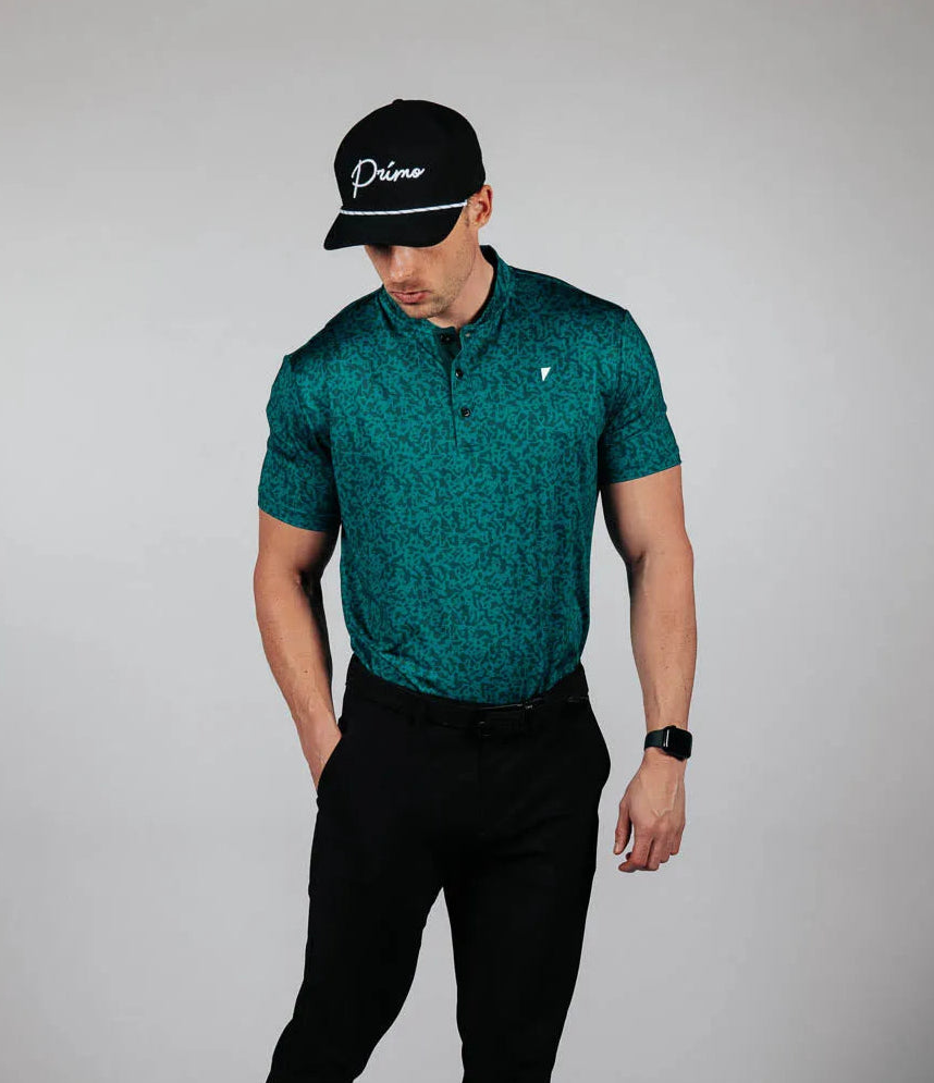 Man wearing a green patterned shirt, black pants, and a black cap on a gray background