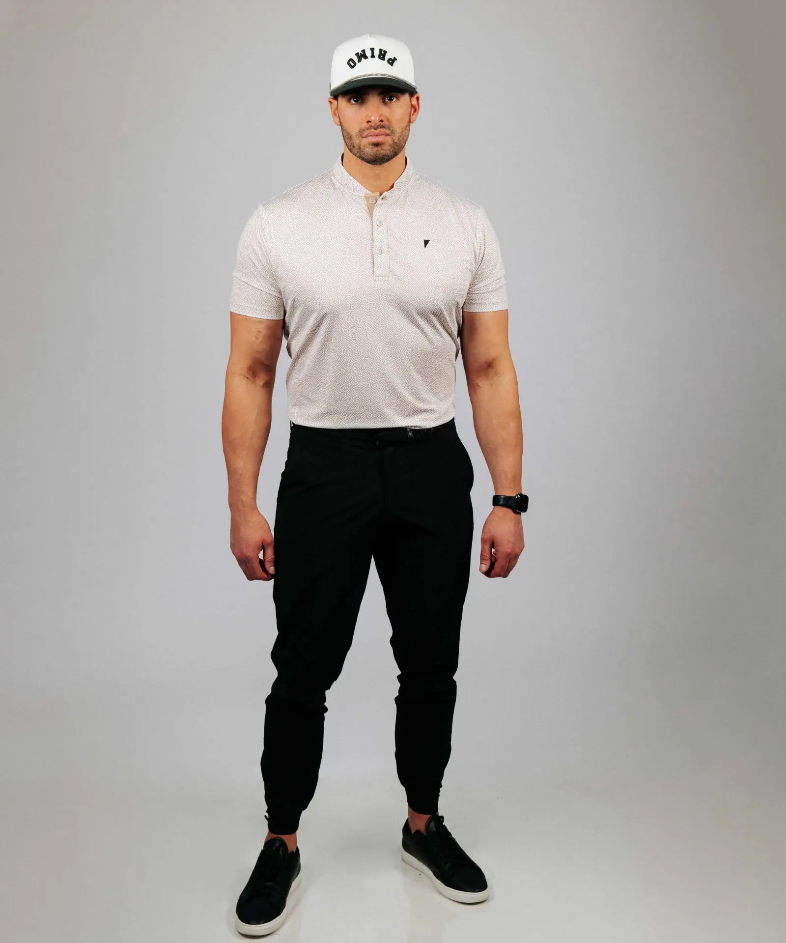 Man wearing a sand coloured polo shirt, black pants, and a white cap with a logo on a grey background