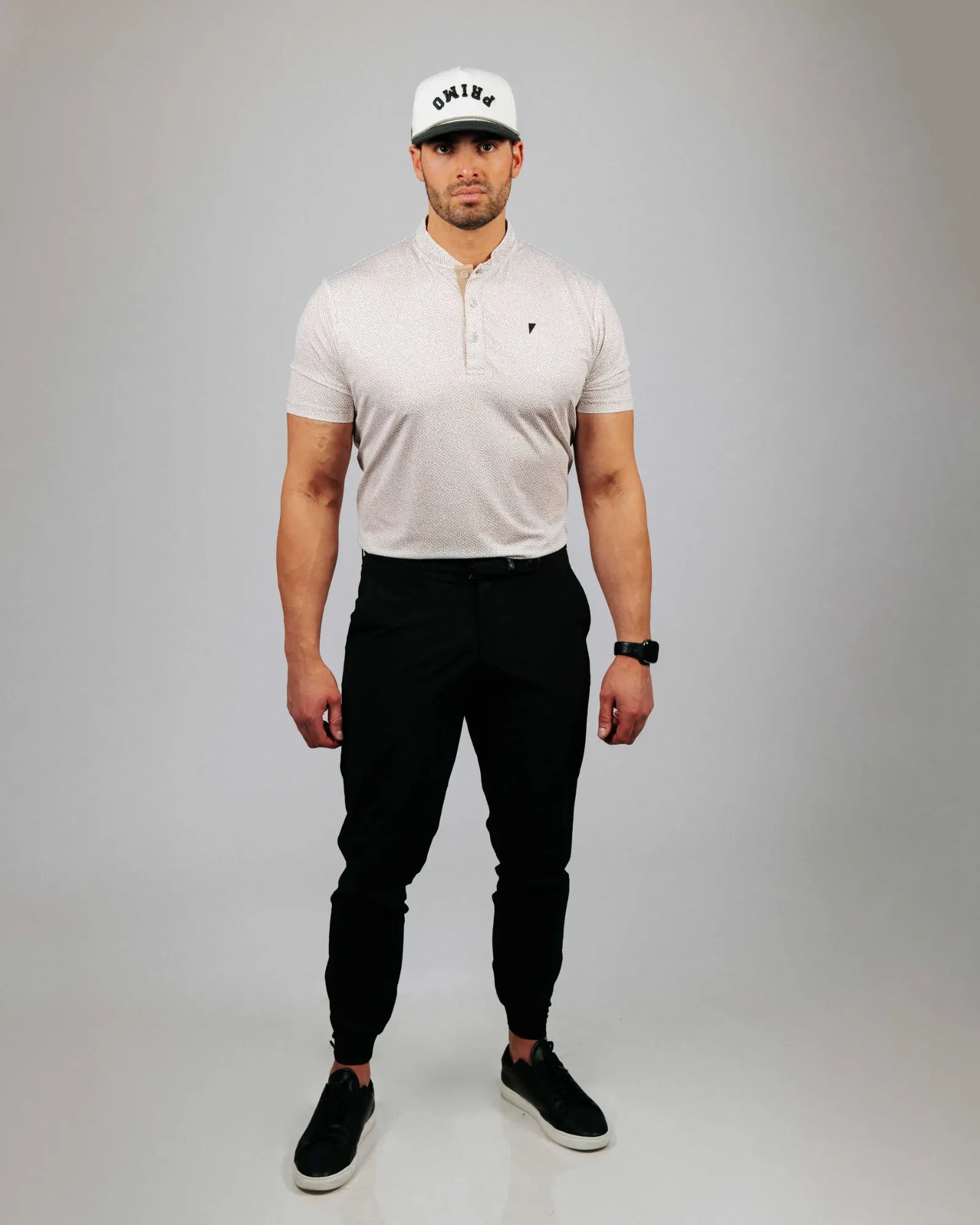 Man wearing a sand coloured polo shirt, black pants, and a white cap with a logo on a grey background