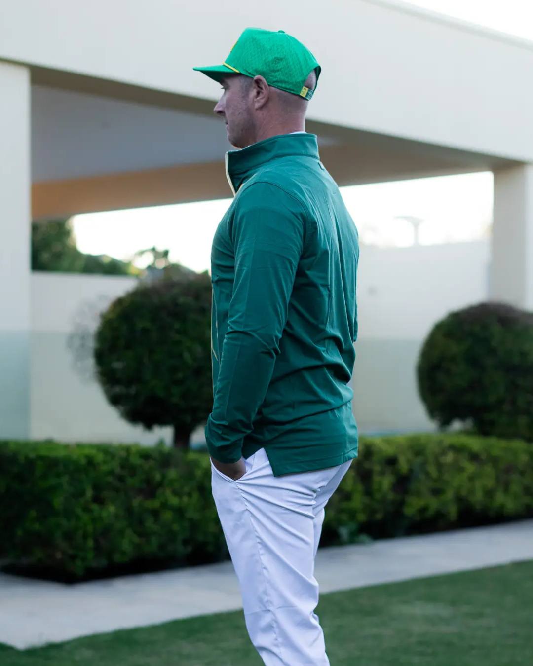 Person wearing a green jacket and cap standing outdoors with greenery in the background