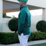 Person wearing a green jacket and cap standing outdoors with greenery in the background