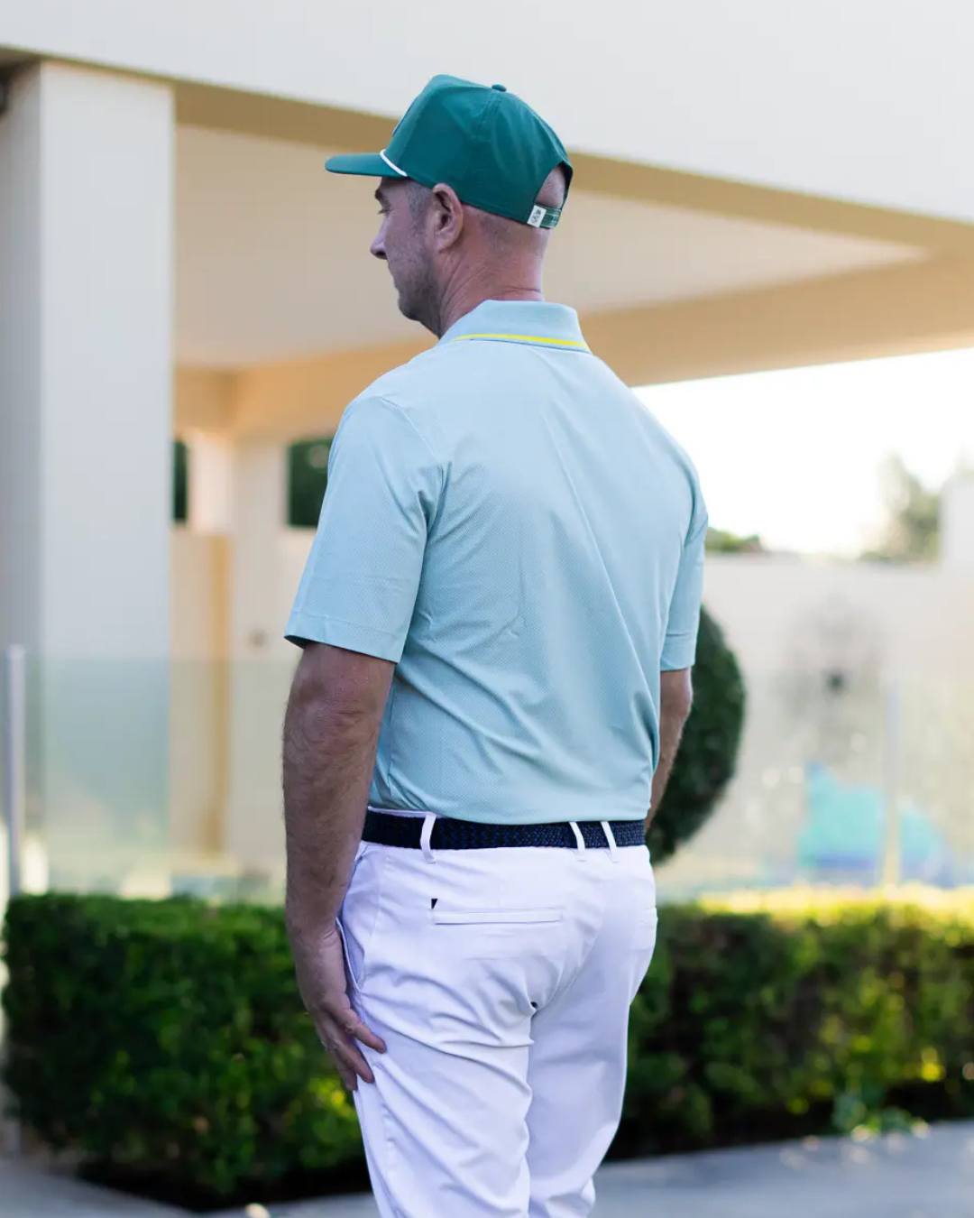 Man wearing a light blue polo shirt and white pants with a green cap, standing outdoors.