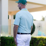 Man wearing a light blue polo shirt and white pants with a green cap, standing outdoors.