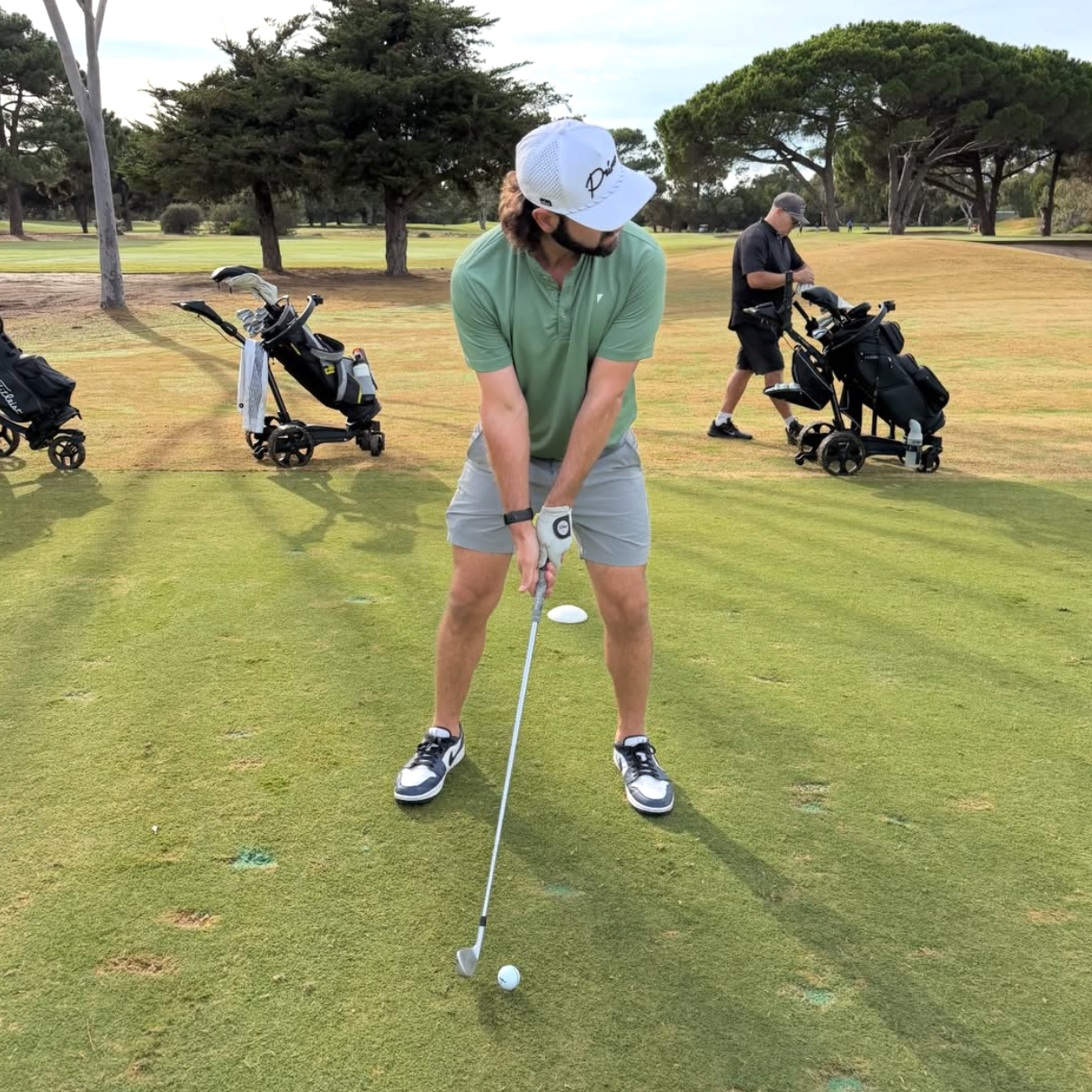 Person playing golf on a green course with trees and other golfers in the background.