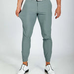 Person wearing smoke green pants with a white shirt and white shoes on a plain background. Model showing how stretchy the pants are.