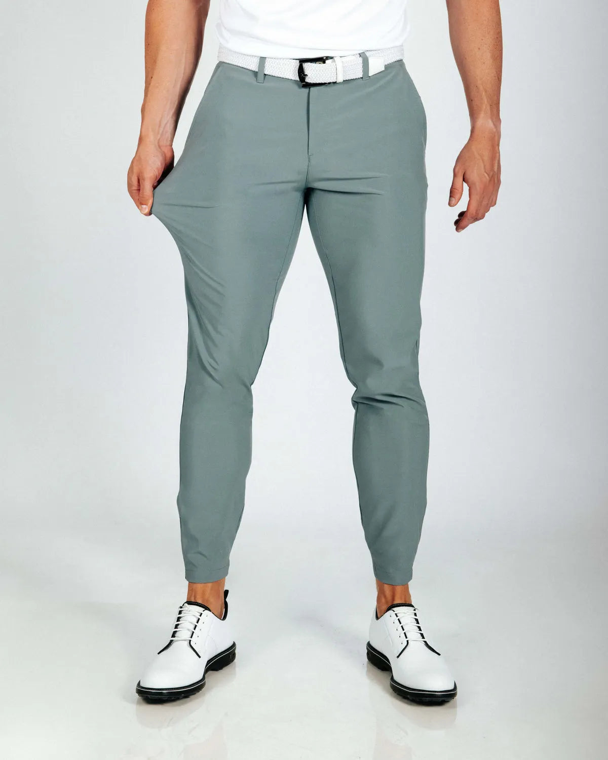 Person wearing smoke green pants with a white shirt and white shoes on a plain background. Model showing how stretchy the pants are.