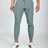 Person wearing smoke green pants with a white shirt and white shoes on a plain background. Model showing how stretchy the pants are.